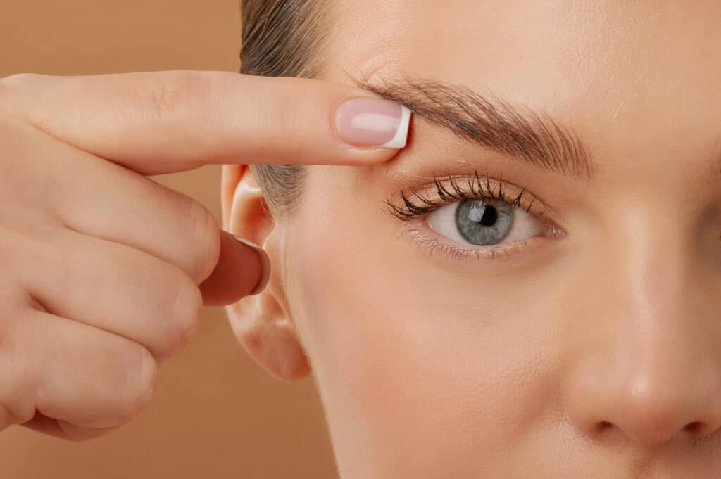 Woman gently touching her upper eyelid, representing the eye area after fully recovering from blepharoplasty. Woman gently touching her upper eyelid, representing the eye area after fully recovering from blepharoplasty.
